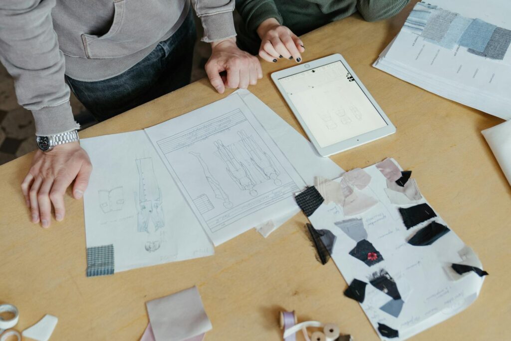 Two designers examine clothing sketches and fabric swatches on a worktable, highlighting fashion creativity.