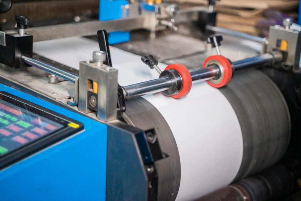 Industrijska mašina sa rolom materijala u procesu štampe etiketa u rolnama, sa metalnim osovinama i crvenim točkićima za vođenje trake.