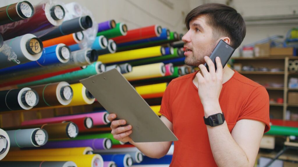 Radnik u štampariji proverava materijale i razgovara telefonom, okružen rolama papira u boji – prikaz procesa kao što je redovna štampa u svakodnevnoj proizvodnji.