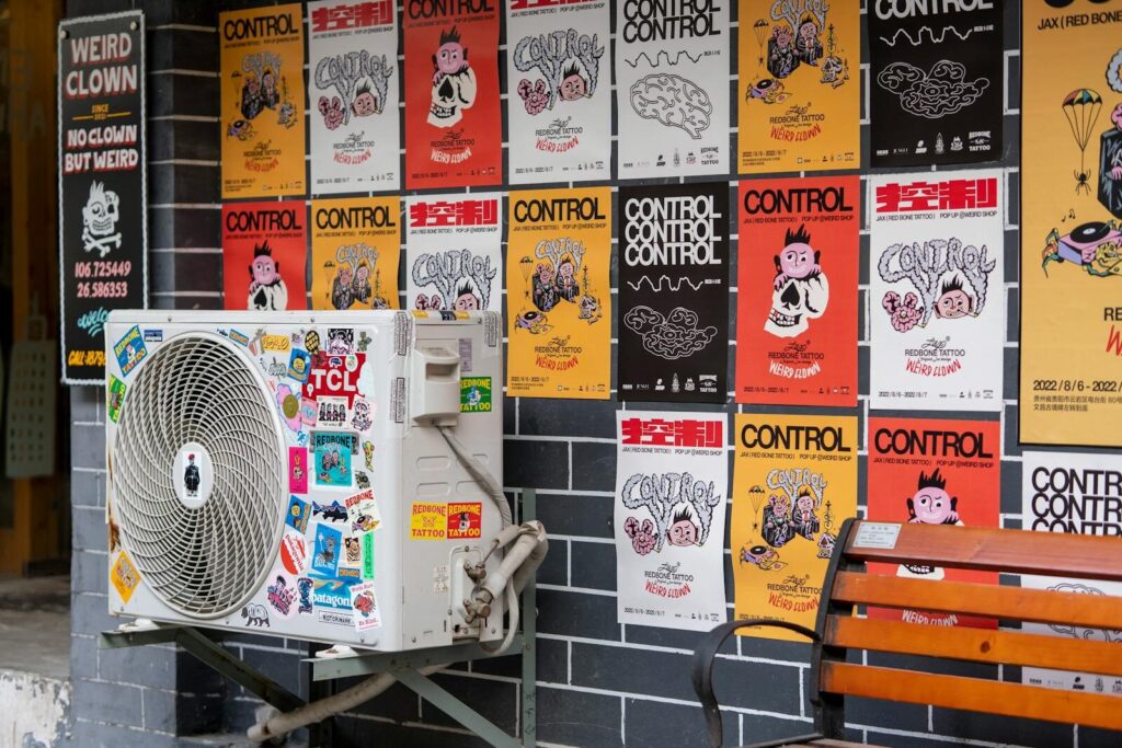 Colorful street art scene featuring a sticker-covered air conditioning unit and bold posters on a brick wall.
