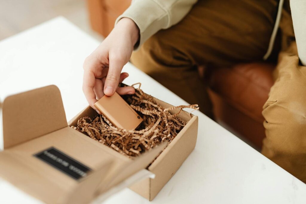 Kratki tiraži ambalaže: kada imaju smisla za male brendove 3 A person unboxes a small item from a cardboard package filled with crinkle paper, indoors.